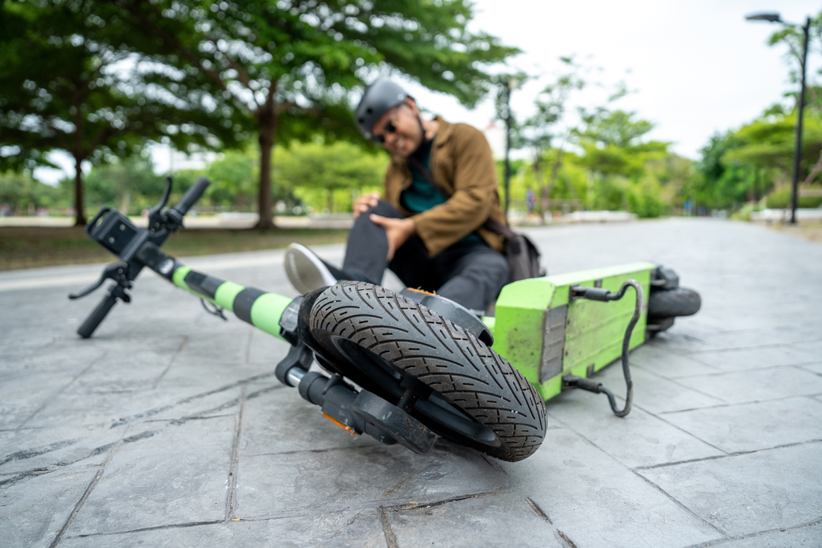 Person wearing a helmet sits on the ground beside a lime-green electric scooter tipped over on a paved park path during daytime.